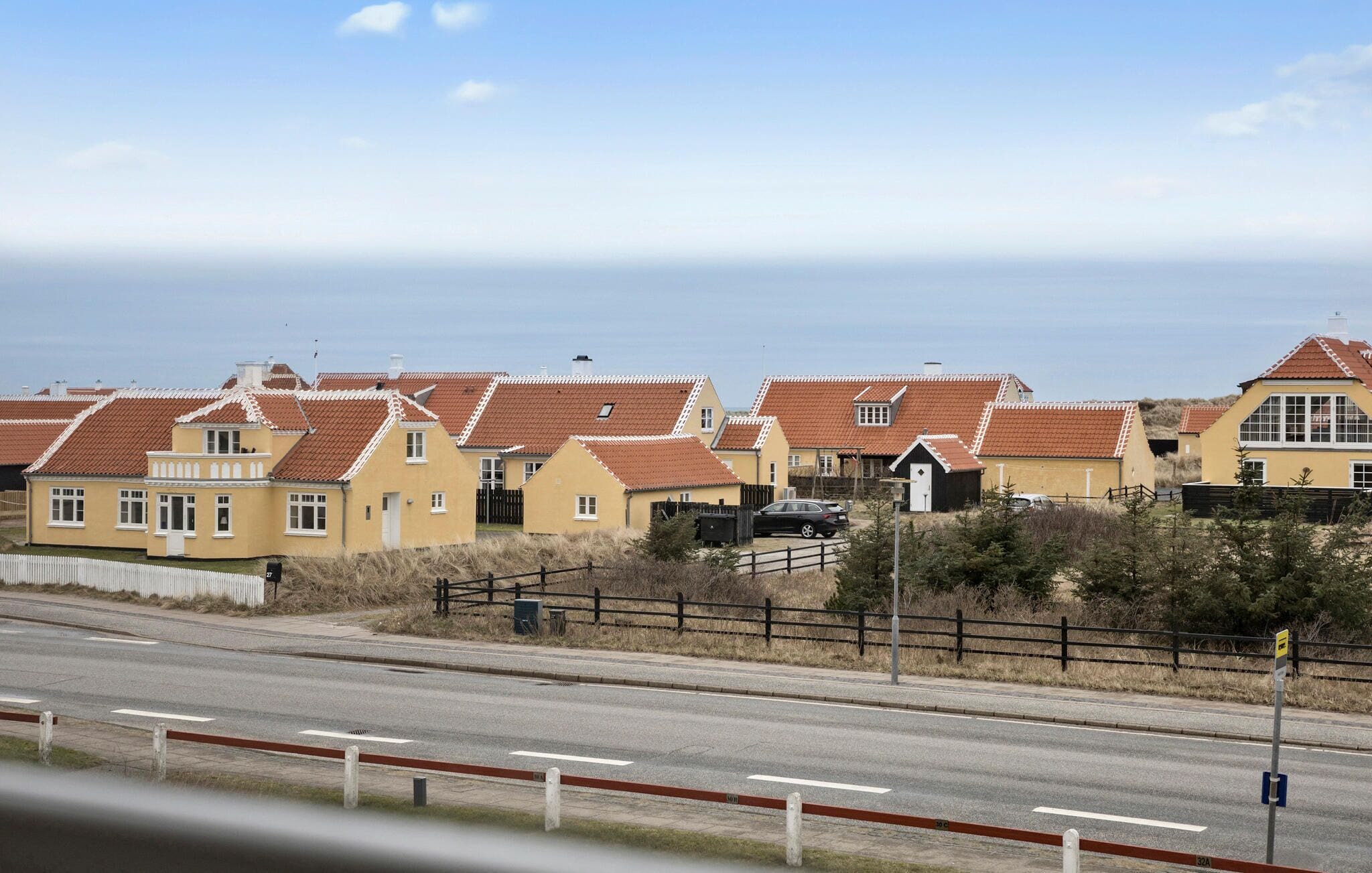 Une zone résidentielle côtière où les maisons se distinguent par leurs toits recouverts de tuiles rouges et leurs murs jaunes, une route avec des marquages blancs délimitant les voies, et une vue dégagée sur la mer en arrière-plan.