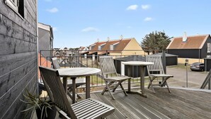 Una terraza de madera con mobiliario exterior, vista a casas vecinas y cielo despejado.