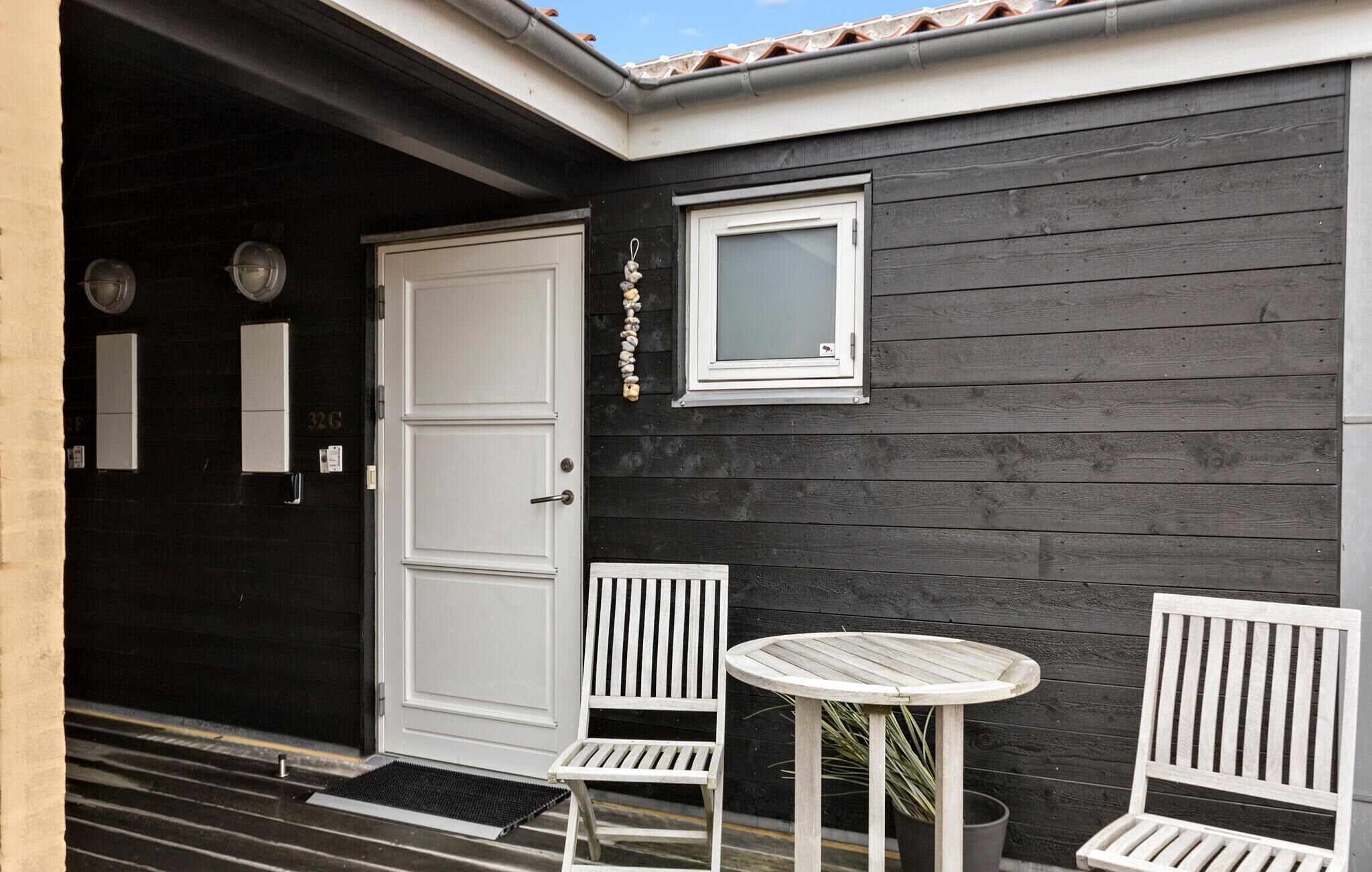 Exterior em madeira preta com uma porta branca, duas cadeiras brancas, uma mesa redonda pequena e uma janela.