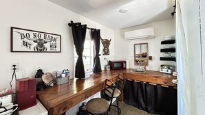 A compact kitchen with a wooden countertop, a microwave, a sink, and a chair. A framed sign is on the wall, and there are shelves with dishes and a decorative item.