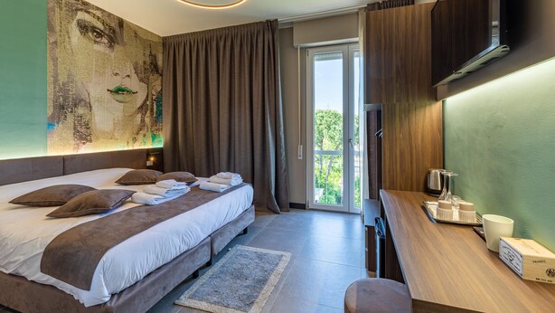 A modern hotel room with a large bed, a desk with a kettle, and a view of the outdoors.