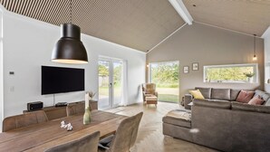 A spacious living room with a large flat-screen TV, a wooden dining table, and a grey sofa.