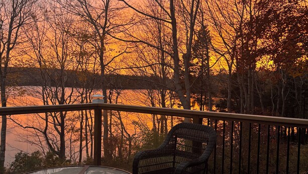 A deck with wicker chairs and a glass table, overlooking a body of water with trees in the background.