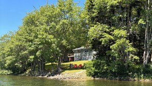 A lakeside cabin with a deck and red chairs, surrounded by dense trees.