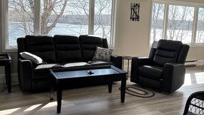 A modern living room with a black leather sofa, a black leather armchair, and a black coffee table. There are large windows with a view of trees and a body of water.