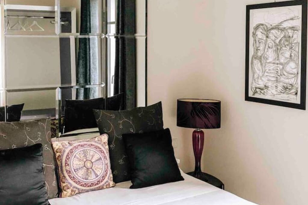 Un lit bien fait, agrémenté d’oreillers noirs et à motifs, une lampe de chevet et une œuvre d’art encadrée au mur.