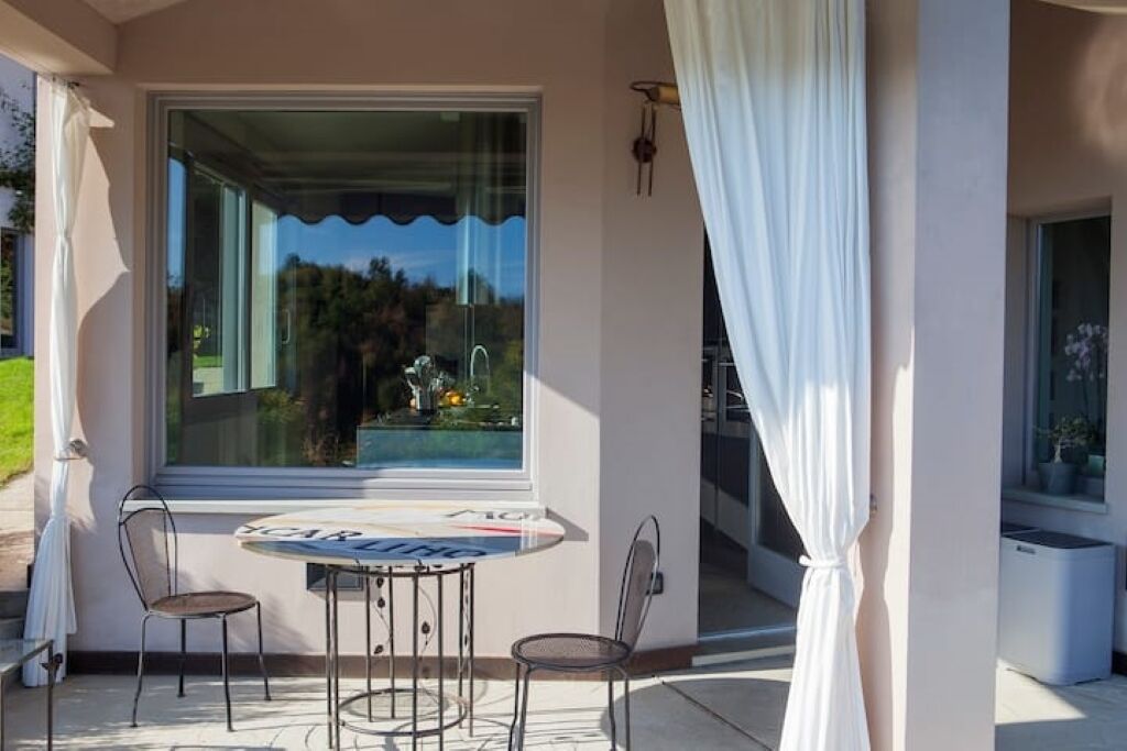 Un balcón con mesa y sillas, una ventana que da a zonas verdes y una cortina blanca.