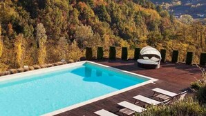 A swimming pool with a wooden deck, lounge chairs, and a cushioned seating area overlooking a forested hillside. - Tenuta Moncarlino - Camera Luce (Piemonte)