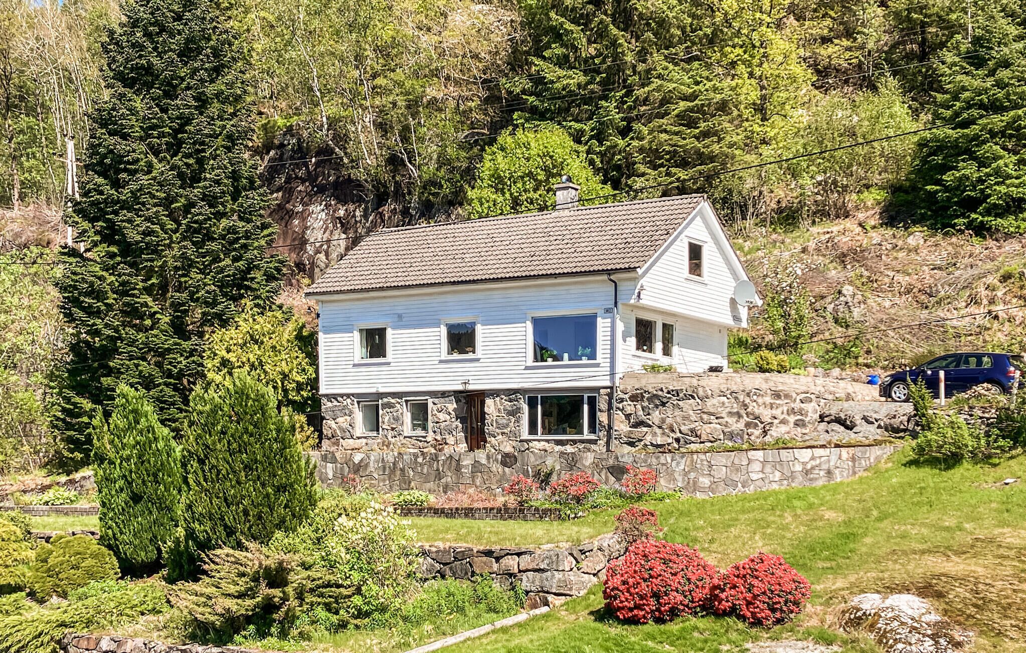 Une maison à deux étages, avec des fondations en pierre, entourée d’une végétation luxuriante et d’un petit jardin agrémenté de fleurs rouges.