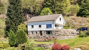 A two-story house with a stone foundation, surrounded by lush greenery and a small garden with red flowers.