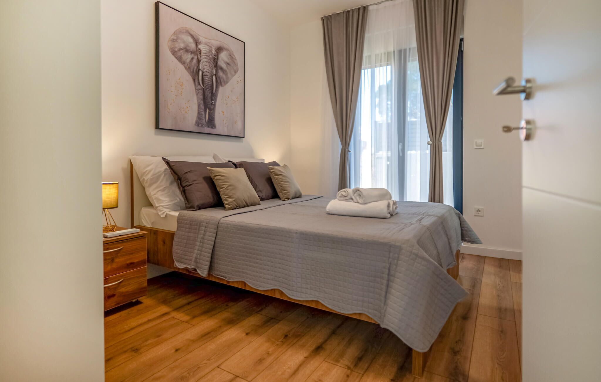A neatly made bed with a grey quilt, a white towel, and several pillows in a modern bedroom with wooden flooring and a large window with curtains.