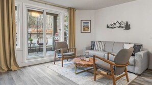 Un salon avec un canapé gris, deux fauteuils, une table basse et une grande porte-fenêtre coulissante donnant sur un espace extérieur.