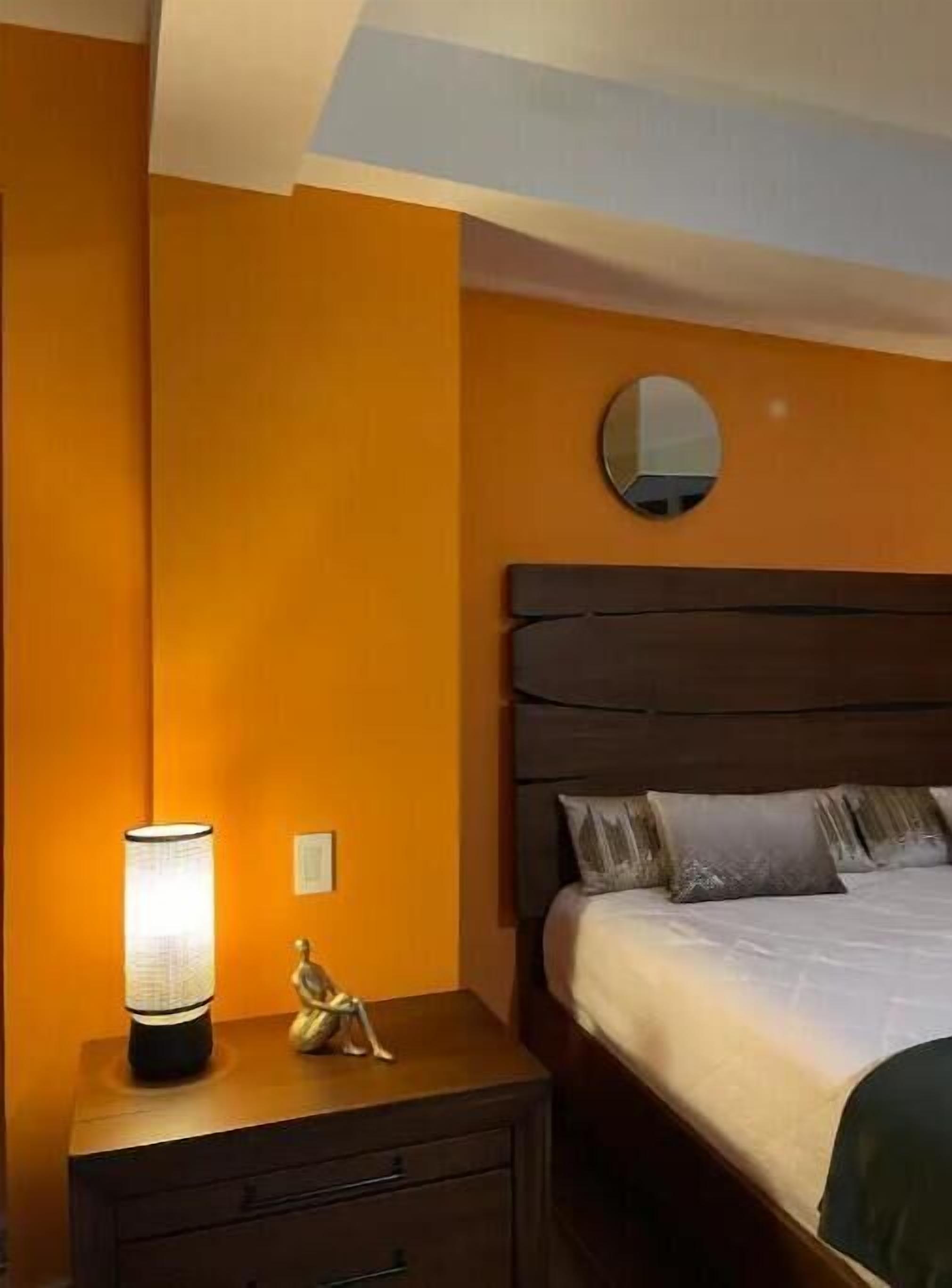 A hotel room with a bed, a nightstand with a lamp, a small statue, and a round mirror on the wall.