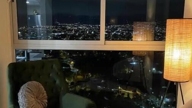 A room with a large window overlooking a cityscape at night, a green armchair with a cushion, and a floor lamp. - Sky View Penthouse Costa Rica (Empalme)