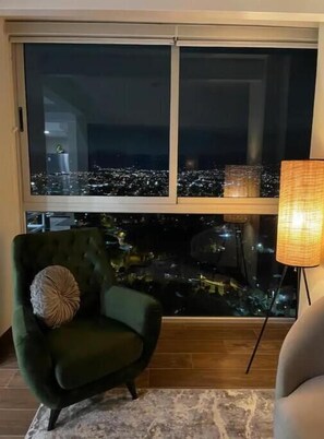 Une pièce dotée d’une grande fenêtre donnant sur une vue nocturne de la ville, d’un fauteuil vert agrémenté d’un coussin et d’un lampadaire.