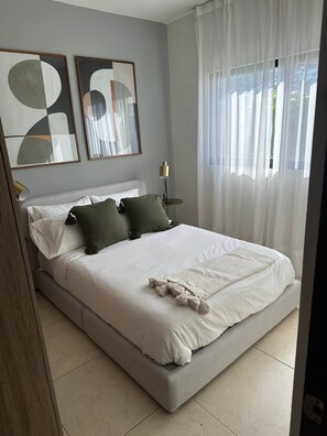 Een slaapkamer met een bed, kussens, een nachtkastje met een lamp, en twee abstracte schilderijen aan de muur.
