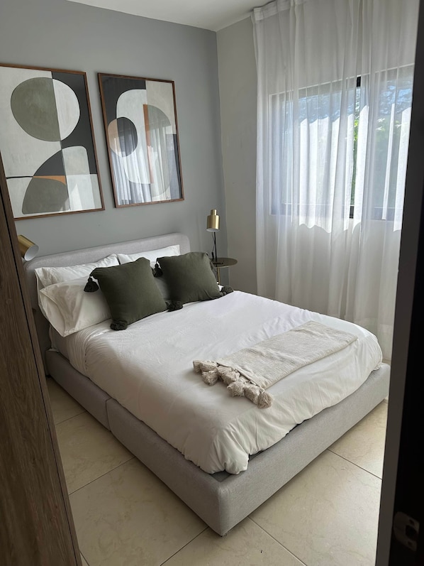 Ein Schlafzimmer mit Bett, Kissen, einem Nachttisch mit Lampe und zwei abstrakten Bildern an der Wand.