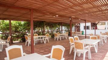 Un patio exterior con mesas y sillas blancas, cubierto por una pérgola de madera.