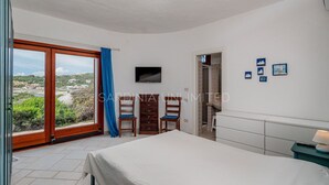 A bedroom with a bed, two chairs, a TV, and a view of the landscape. - Villa Lady Oceania - Sardinia Unlimited (Santa Reparata)