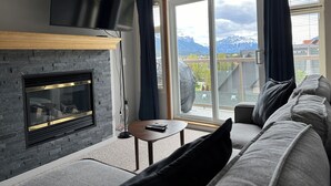 Una sala de estar moderna con chimenea de piedra, un televisor de pantalla plana montado sobre ella, un sofá y vistas a las montañas a través de amplias ventanas.