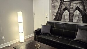 A living room with a dark wooden coffee table, a black leather sofa, a wall-mounted lamp, and a large painting of a bridge. - Deluxe! New Apt Loft Near Downtown – Ready for Families and Business Travelers. (Raleigh)