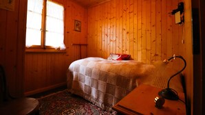 A cozy room with wooden walls, a bed with a checkered blanket, a bedside lamp, and a window with curtains. - Charming detached cottage for up to 5 people. It has a good location close to a shuttle bus stop tha (St-Luc)