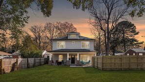 A two-story house with a front porch, a fenced backyard, and trees surrounding the property. - Epic Group Stay: 2 Homes + Hot Tubs + Yard Games (Charlotte)
