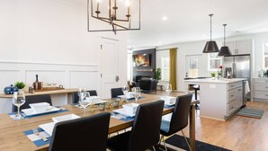 A modern kitchen with a central island, a dining area with a wooden table and chairs, and a living area with a fireplace. - Epic Group Stay: 2 Homes + Hot Tubs + Yard Games (Charlotte)
