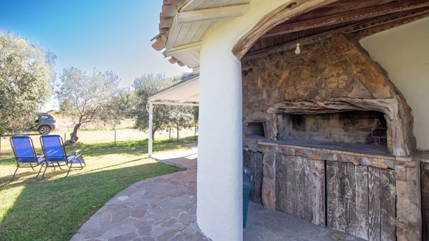 An outdoor patio with a stone fireplace, a covered seating area, and a car parked in the background. - Su Tuvulu 2 by All4sardinia  (CARDEDU)
