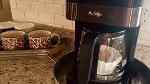 En kaffebryggare med texten ”Mr. Coffee” står på en köksbänk, och bakom den finns en väggbeklädnad av kakel samt två muggar med blommönster.