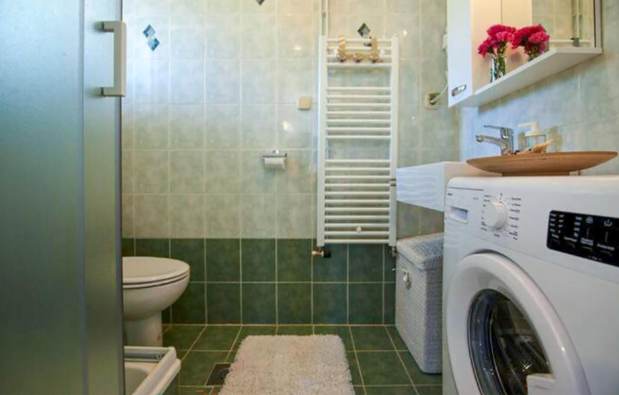 A compact bathroom with a toilet, a washing machine, and a shelf with flowers.