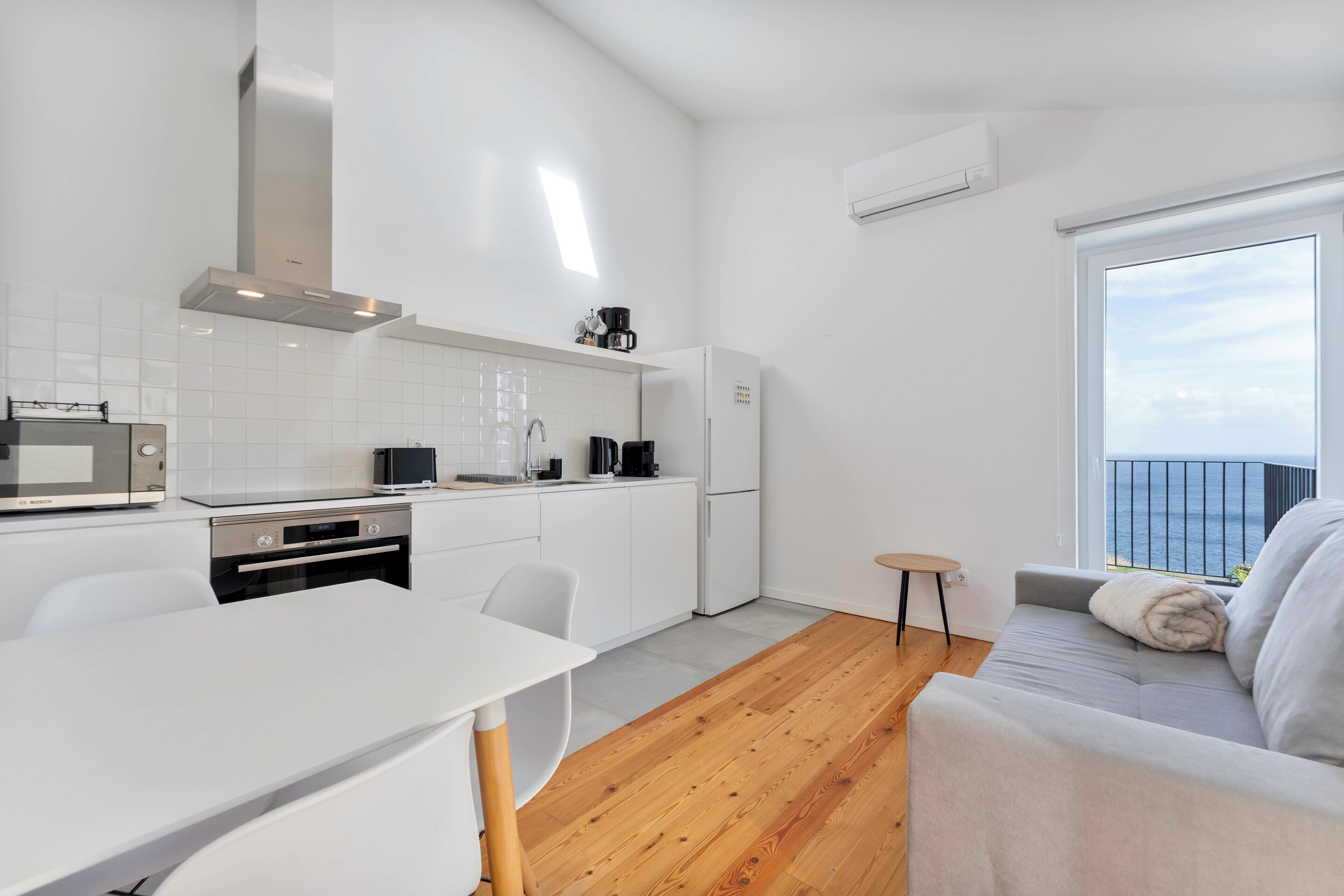 Eine moderne Küche mit weißen Schränken, einem Esstisch und einer Couch an einem großen Fenster mit Meerblick.