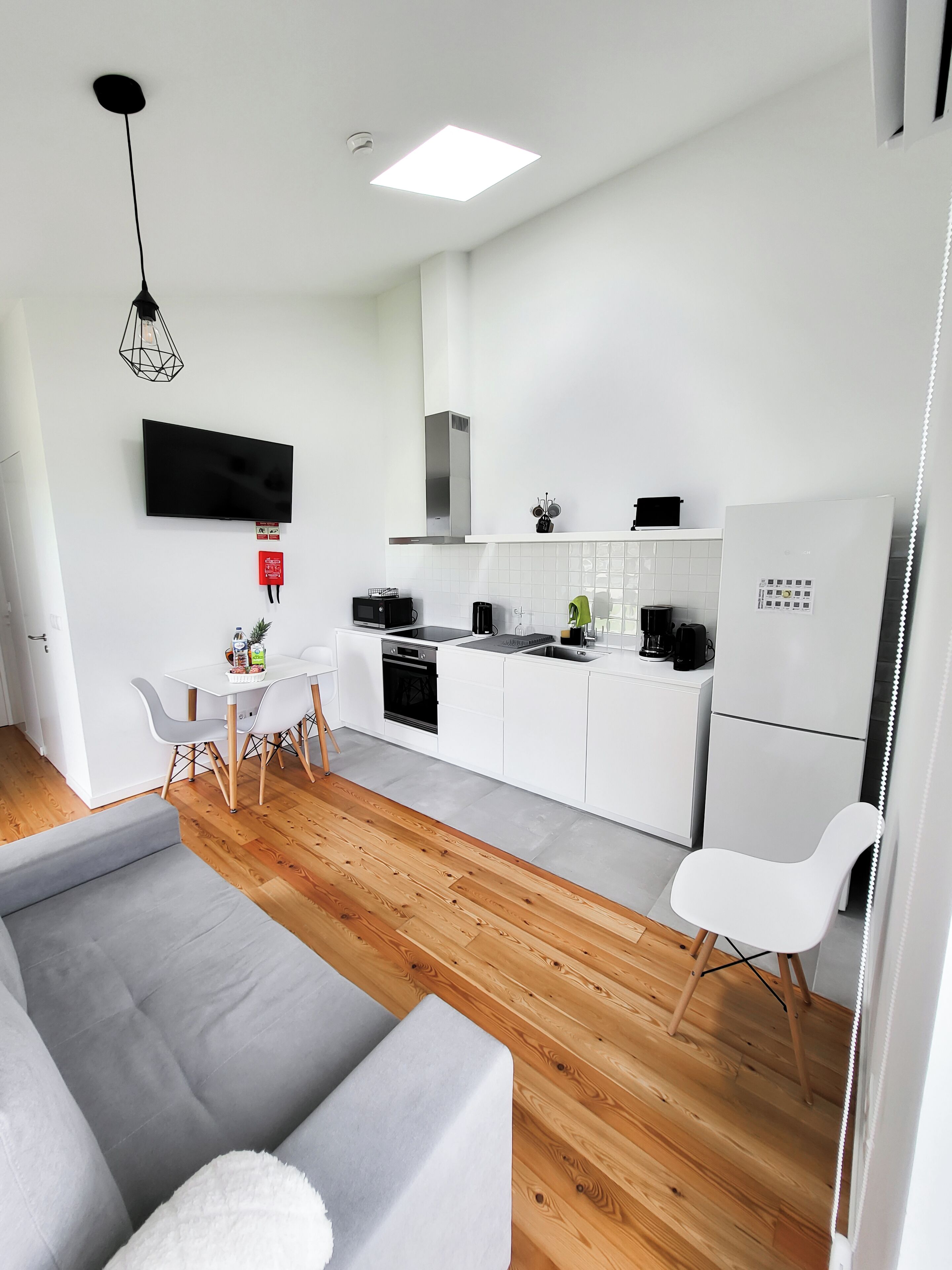 Et moderne køkken med hvide skabe, en indbygget ovn og et køleskab. Der er et spiseområde med en grå sofa og et lille bord.