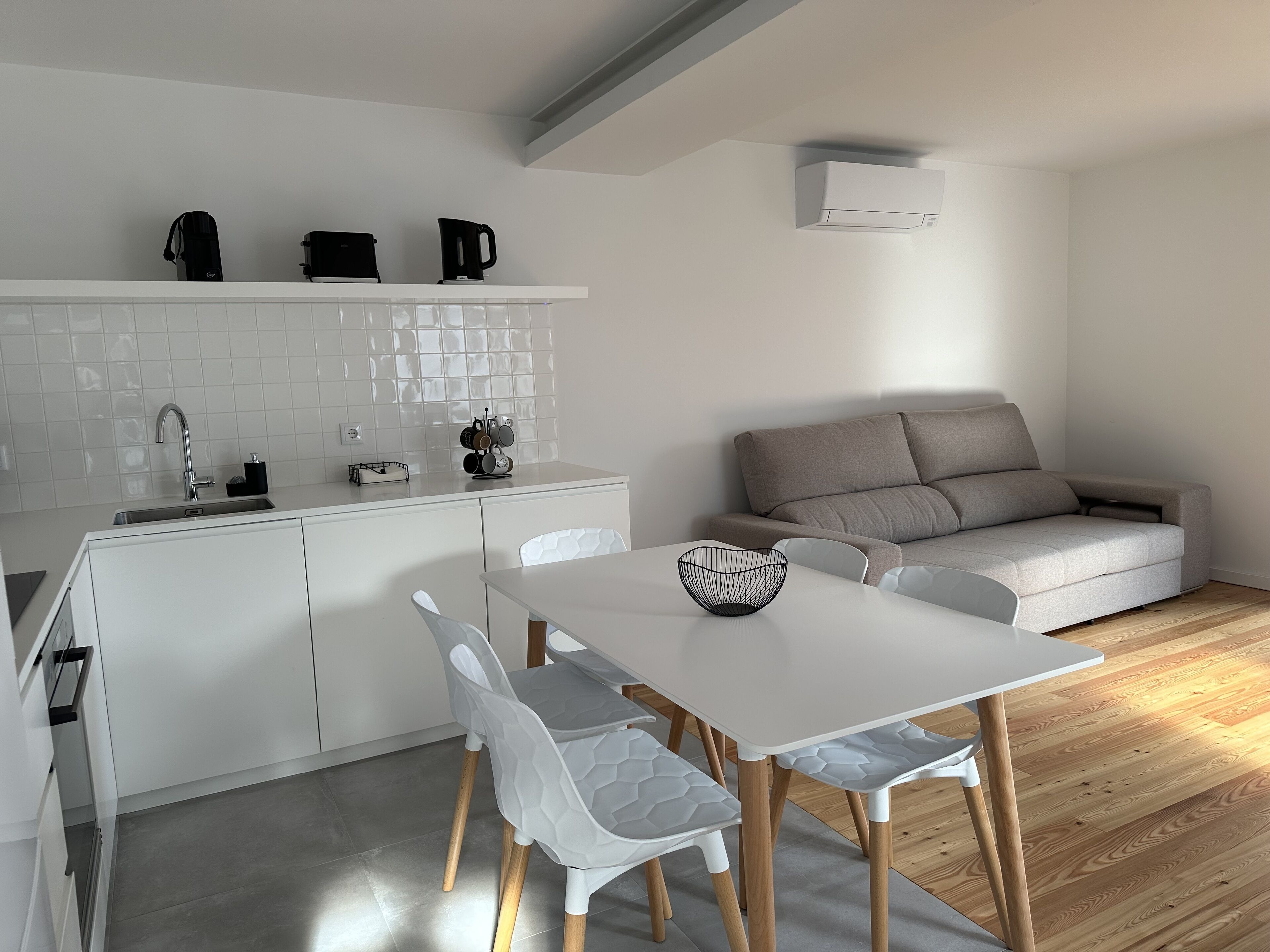 A modern kitchen with white cabinets, a white countertop, and a built-in sink. There is a dining area with a white table and chairs, and a grey sofa in the background.