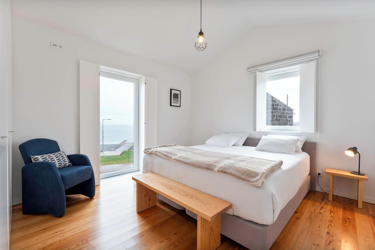 Quarto com cama, banco, cadeira, mesa de cabeceira e vista para o exterior.
