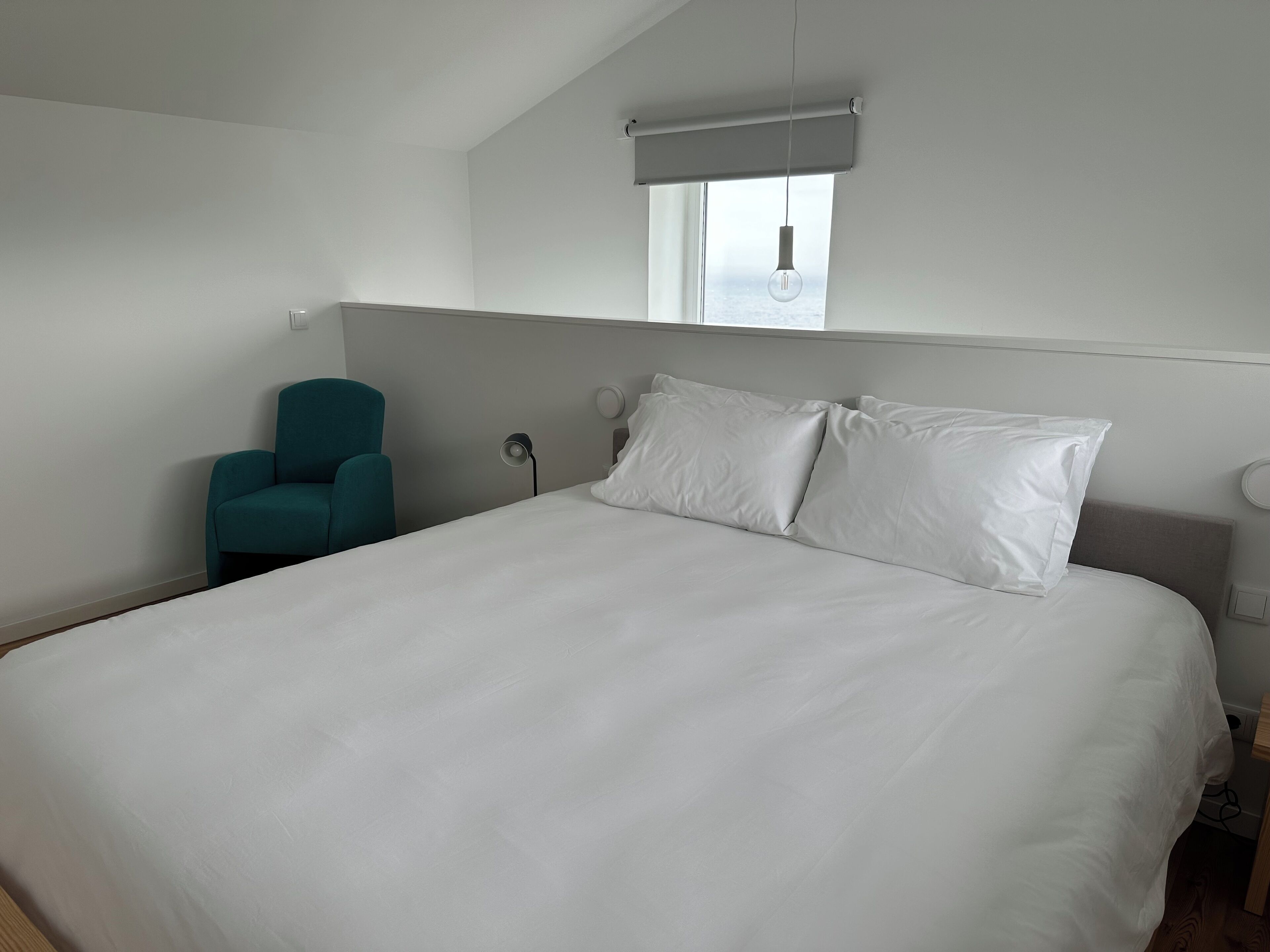 Ein Schlafzimmer mit einem großen Bett, einem türkisfarbenen Sessel, einem Nachttisch, einer Lampe und einem Fenster mit Blick.