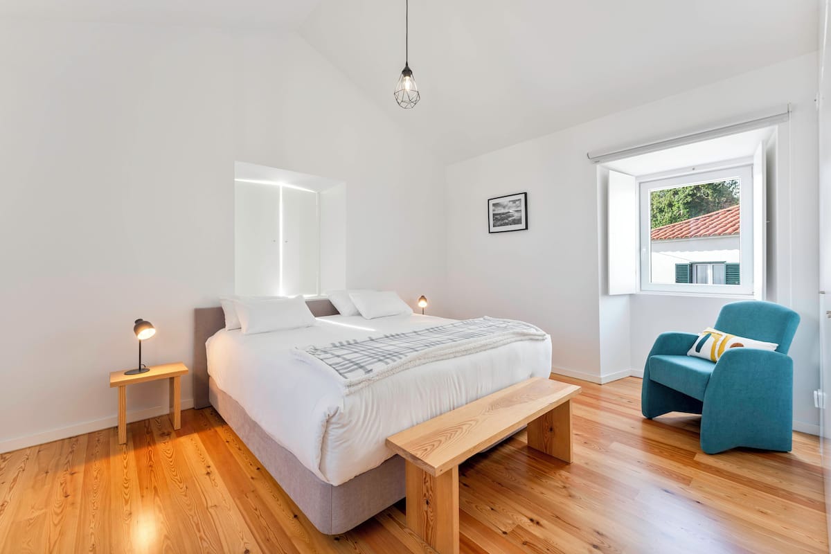 Quarto moderno com uma cama grande, mesas de cabeceira, um banco, uma cadeira e uma janela com vista.