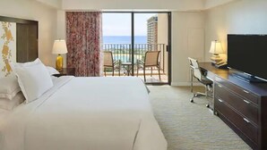 A hotel room with a bed, a desk, a TV, a balcony with a view of the ocean, and a chair. - Waikiki | Couples Surf Nest | Pool + Spa (Honolulu)