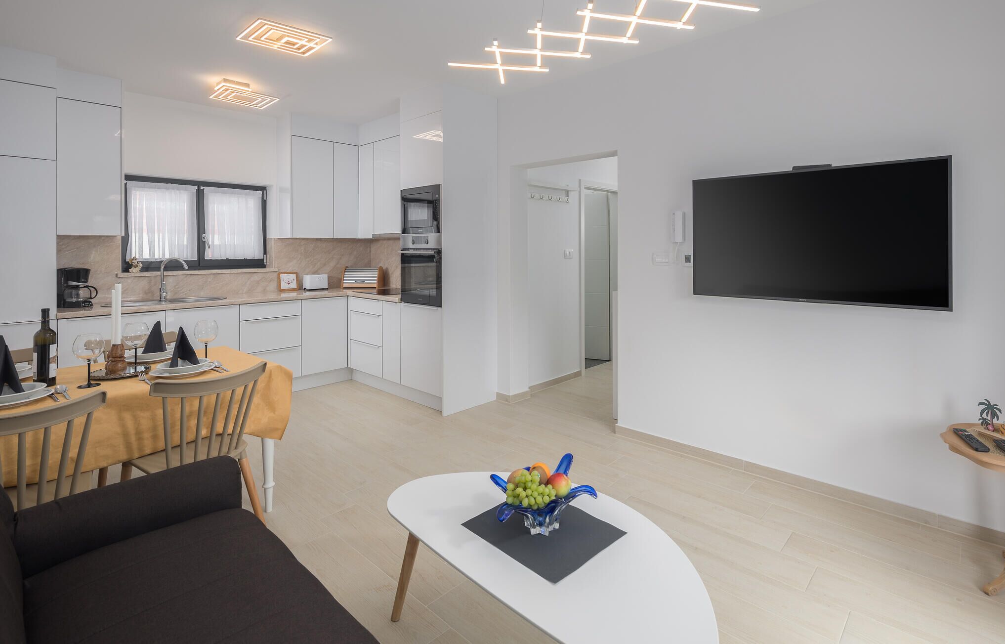 Eine moderne Küche mit weißen Schränken, ein Essbereich mit Tisch für zwei Personen und ein flachbildfernseher an der Wand.
