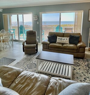Sala de estar com sofá, poltrona e mesa de centro, varanda com cadeiras e um relógio na parede.