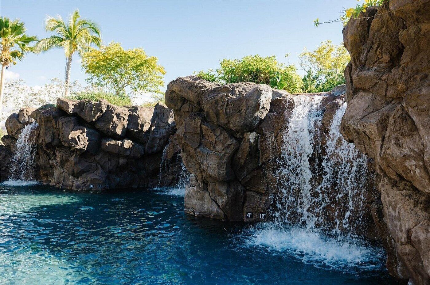 A natural rock formation with a waterfall, clear blue water, and palm trees in the background.