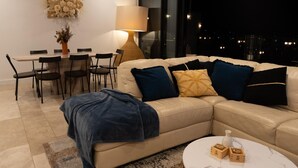 Una sala moderna con un sofá beige, una mesa de comedor con sillas, una lámpara y vistas de la ciudad de noche.