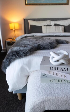 Un lit bien fait, agrémenté d’un plaid en fausse fourrure grise, d’un oreiller blanc et d’une couverture blanche. Un livre de développement personnel intitulé « RÊVE, CROIS, RÉALISE » est posé sur le lit.