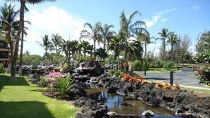 A landscaped area with a small water feature, palm trees, and a paved walkway. - Waikoloa Big Island | Kona Coast Adventure | Pool & Tennis | 2BR (Waikoloa Village)