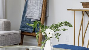 Ett rum med ett glasbord, en vit vas med blommor, ett trästaffli med ett mönstrat tyg och en tv på väggen.