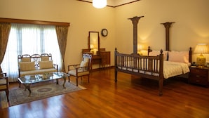 Een ruime kamer met houten vloer, een glazen salontafel, een bank, een stoel, een bed met houten hoofdeinde en een nachtkastje met lamp.