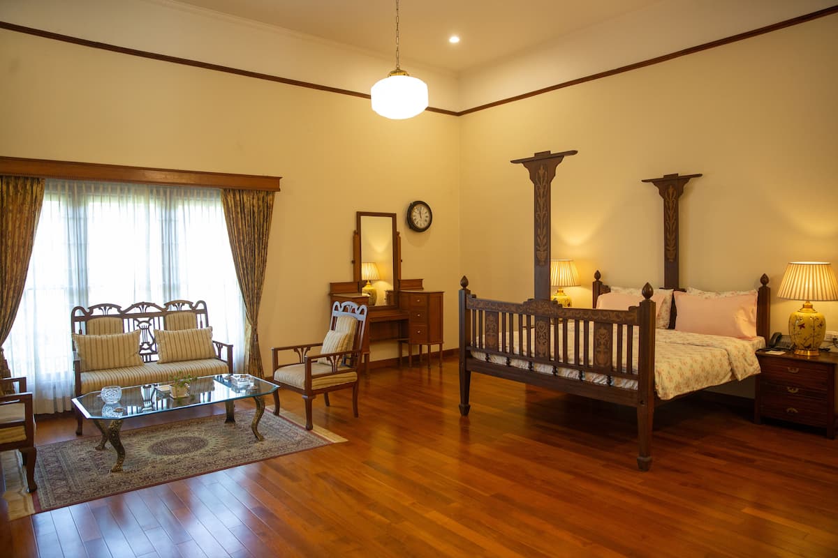 Een ruime kamer met houten vloer, een glazen salontafel, een bank, een stoel, een bed met houten hoofdeinde en een nachtkastje met lamp.