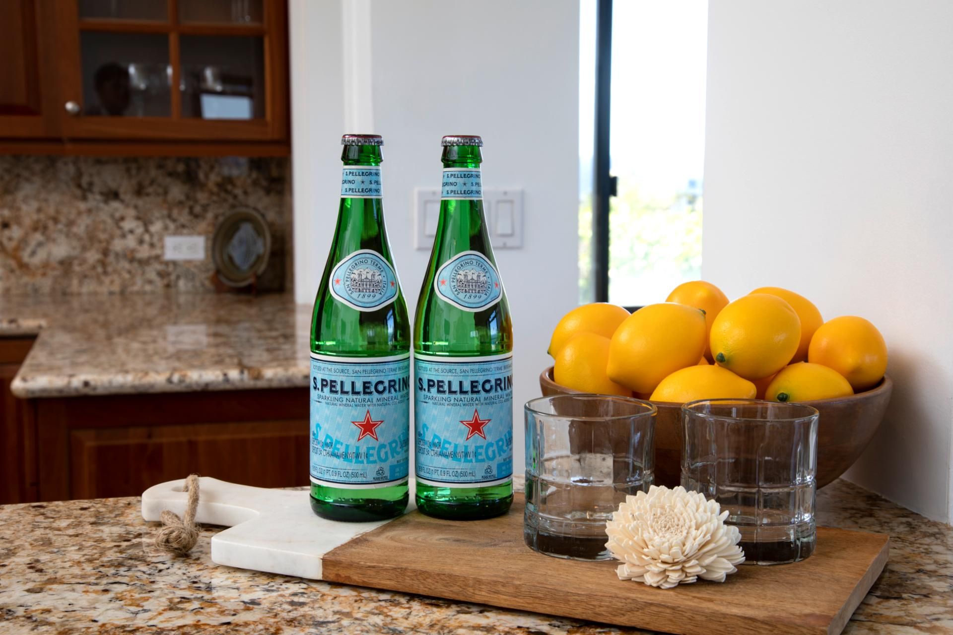 Due bottiglie di acqua minerale frizzante San Pellegrino, una ciotola di limoni e un tagliere di legno con un coltello e un fiore.