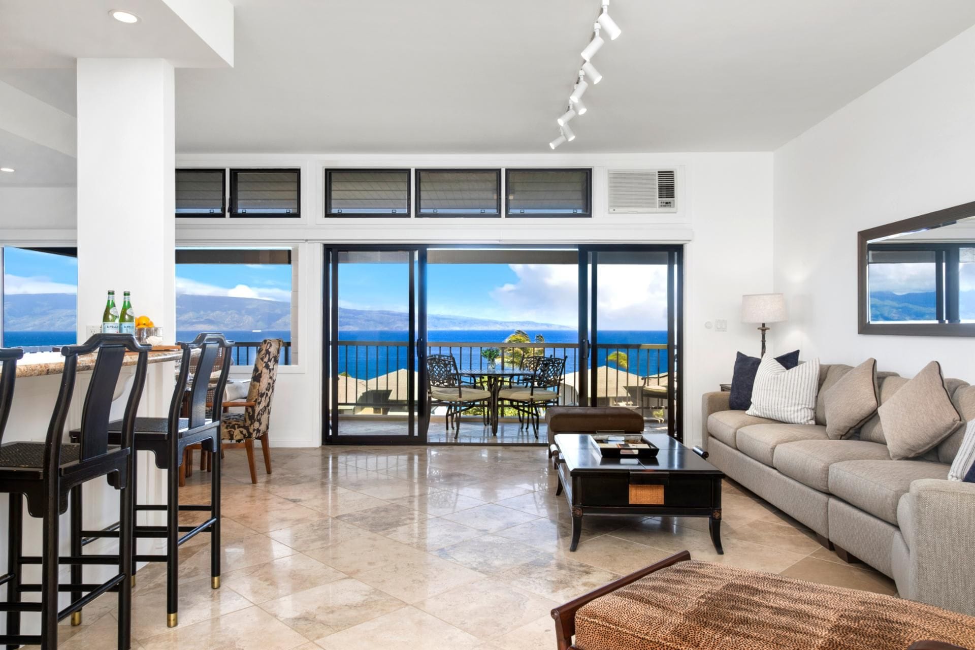 A spacious living area with a large sliding glass door leading to a balcony with a sea view. There is a seating area with a sofa and chairs, a coffee table, and a small table with bottles and a lemon.