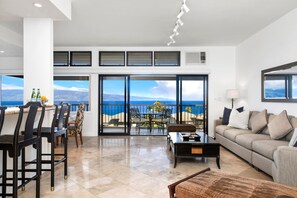 A spacious living area with a large sliding glass door leading to a balcony with a sea view. There is a seating area with a sofa and chairs, a coffee table, and a small table with bottles and a lemon.